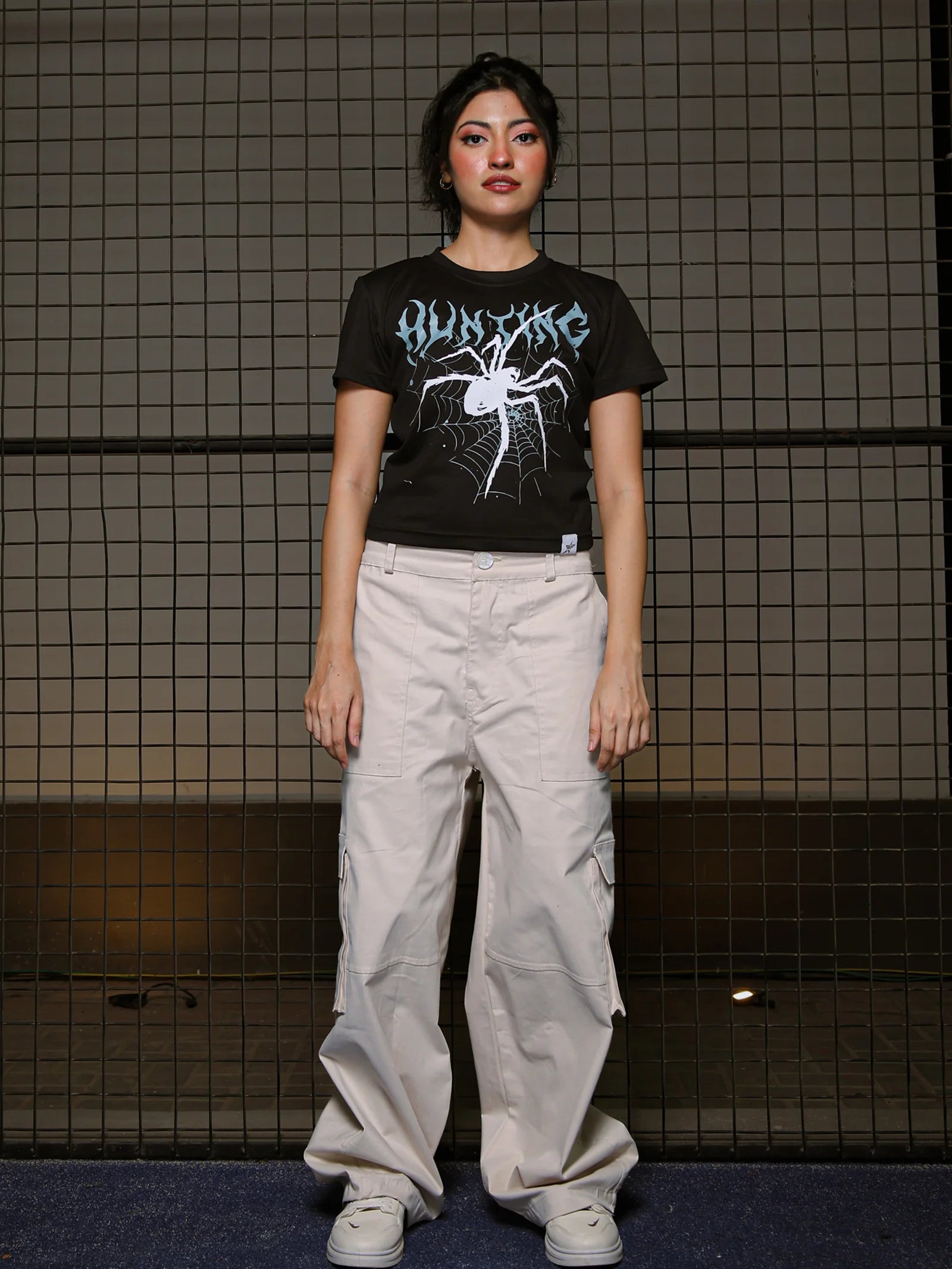 Person wearing a black t-shirt with a graphic design and light-colored pants standing against a tiled wall.