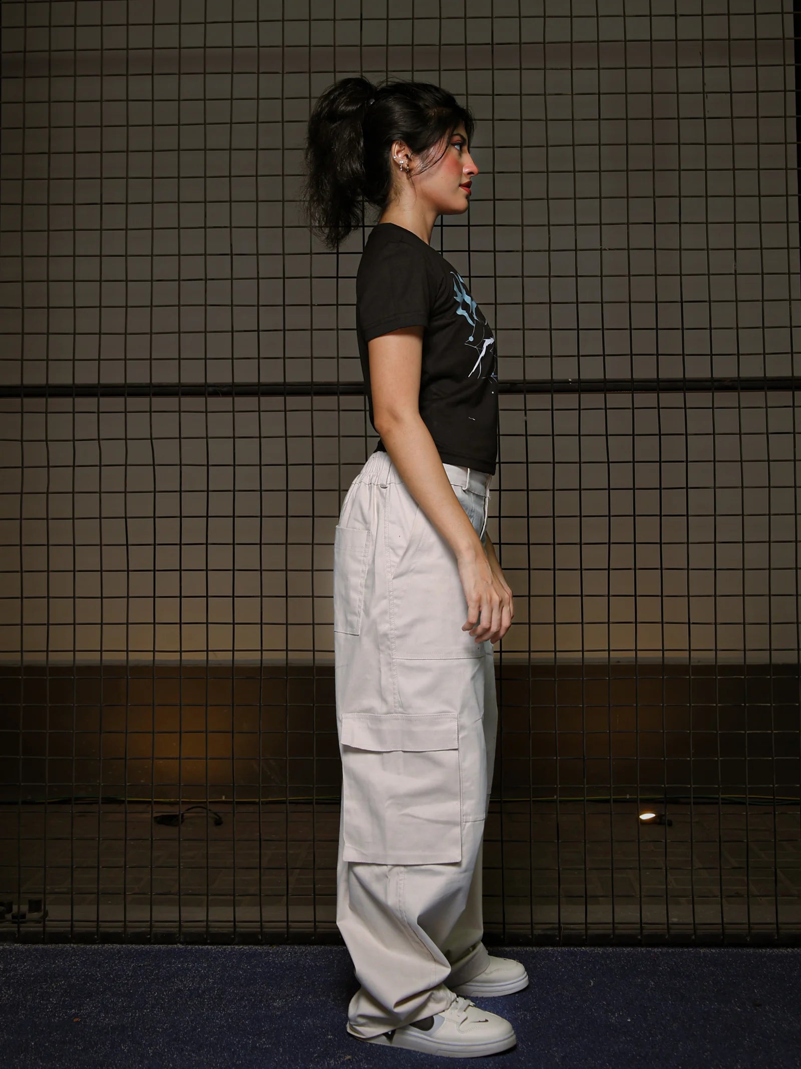 Person wearing a black t-shirt and light gray pants standing against a tiled wall.