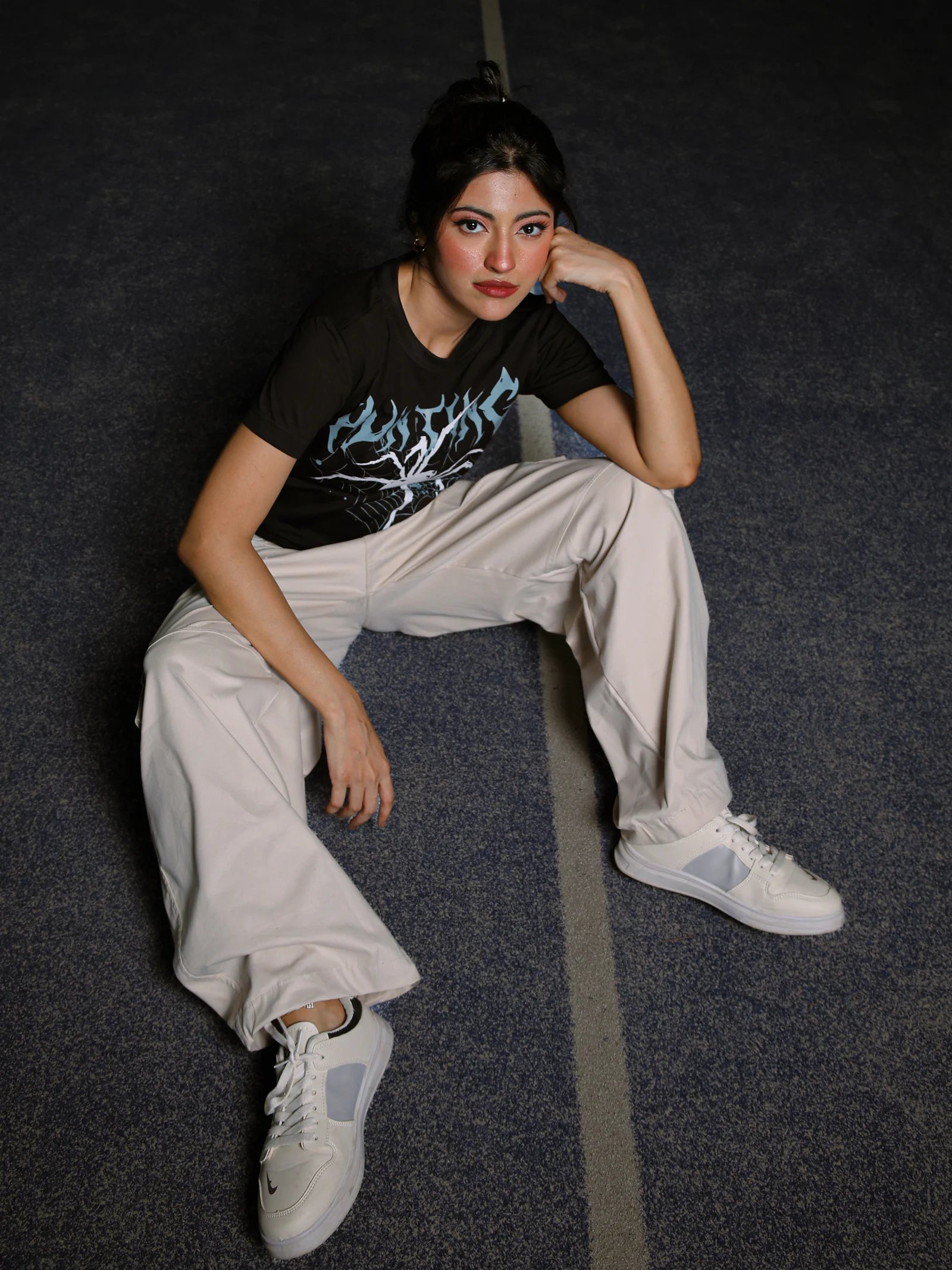 Person sitting on a dark floor wearing a black t-shirt with graphic design, light-colored pants, and white sneakers.