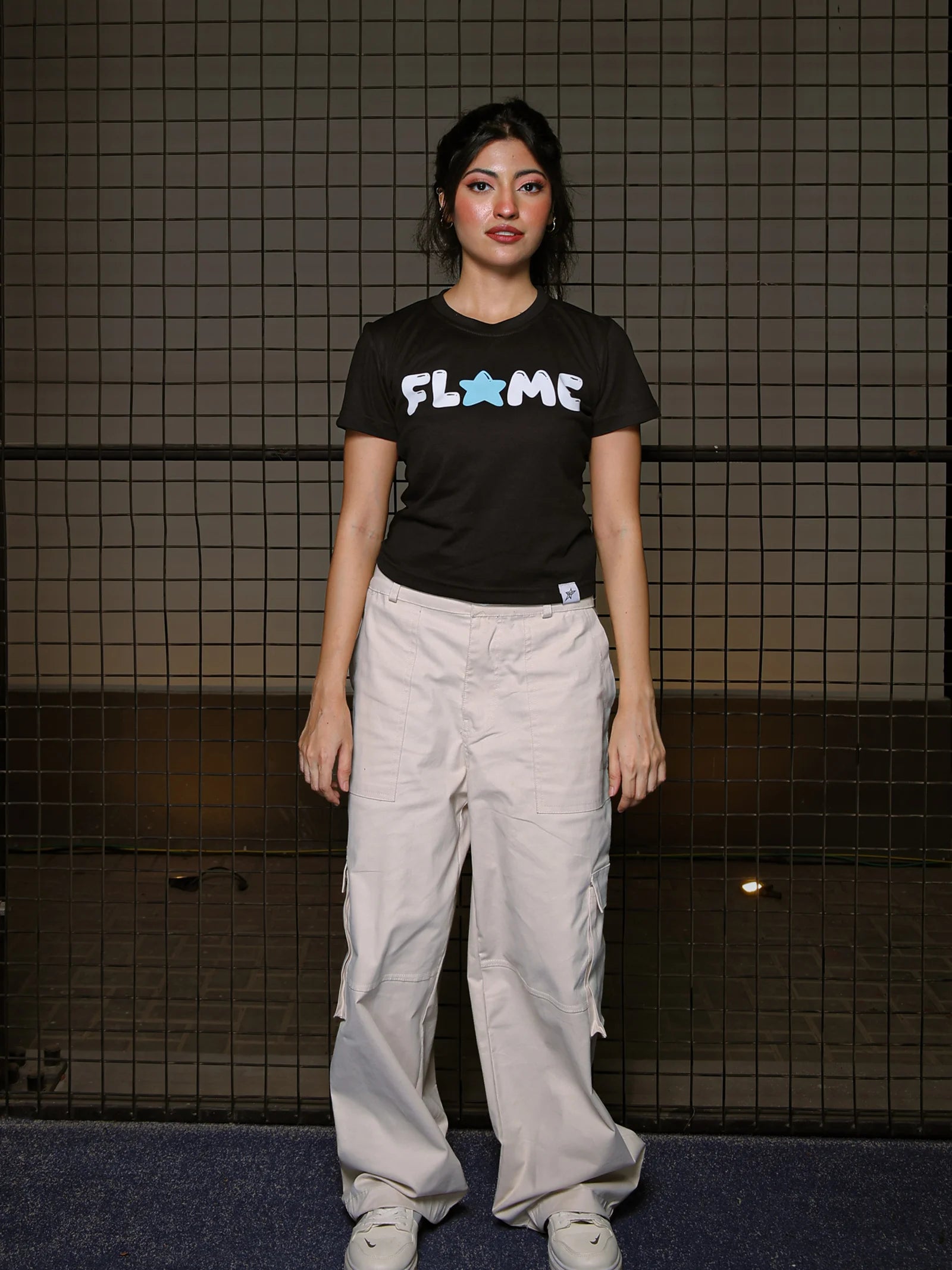 Person wearing a black t-shirt with 'FLAME' logo and light gray pants standing against a dark tiled wall.