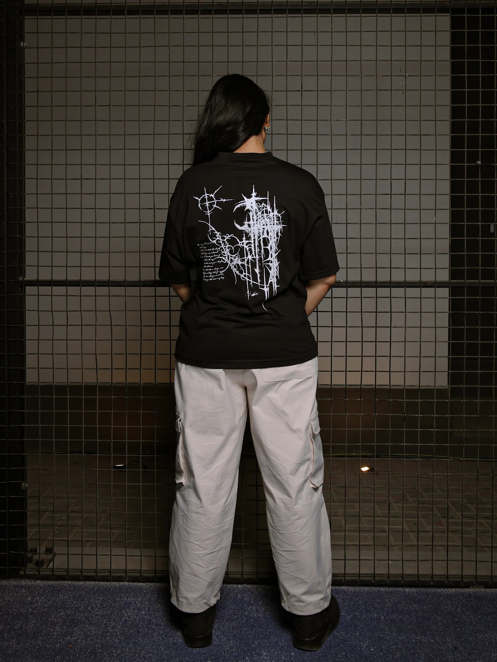 Person wearing a black t-shirt with white graphics and light-colored pants standing against a tiled wall.