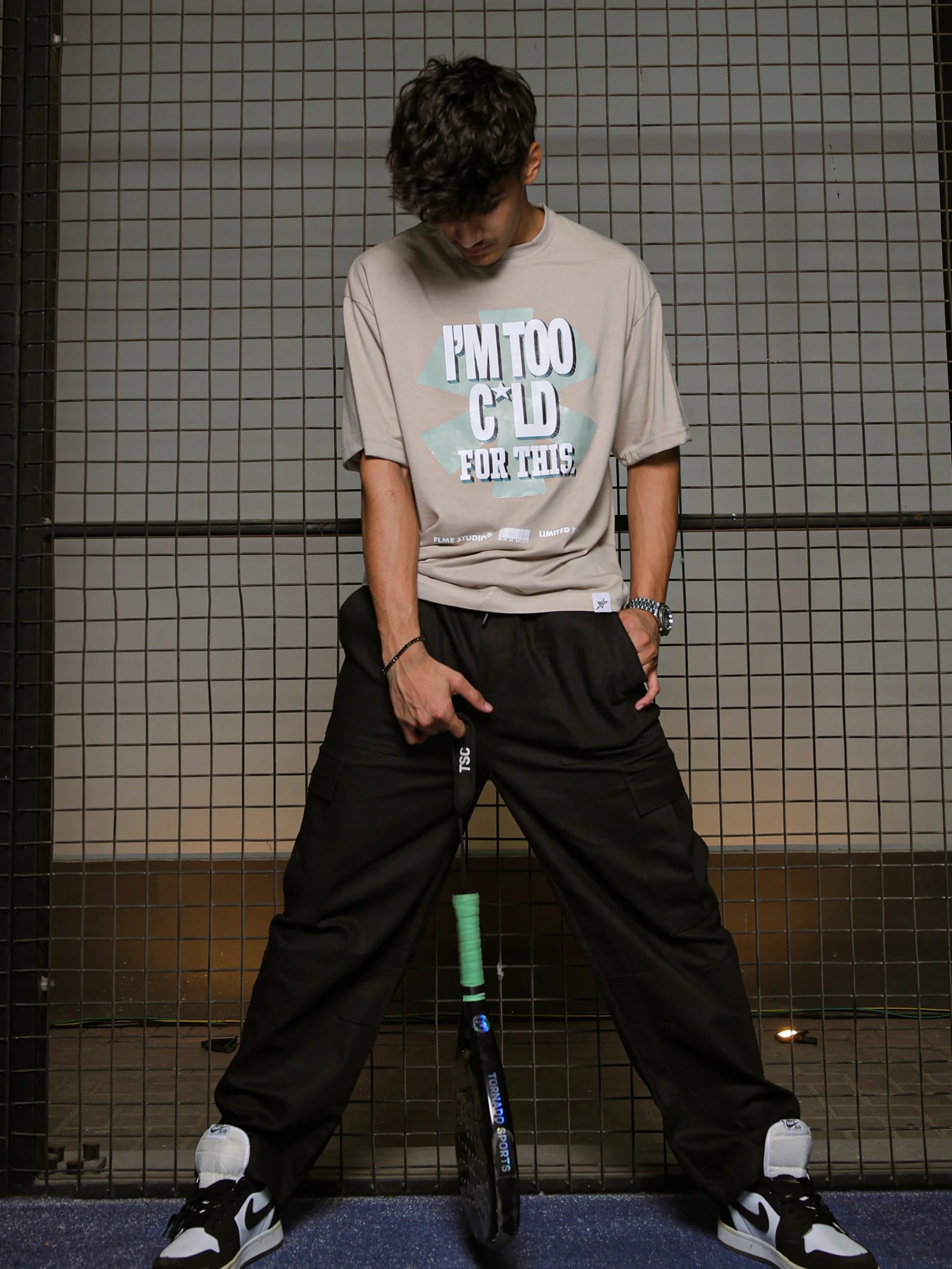 Person wearing a beige t-shirt with text and dark pants, standing against a tiled wall.