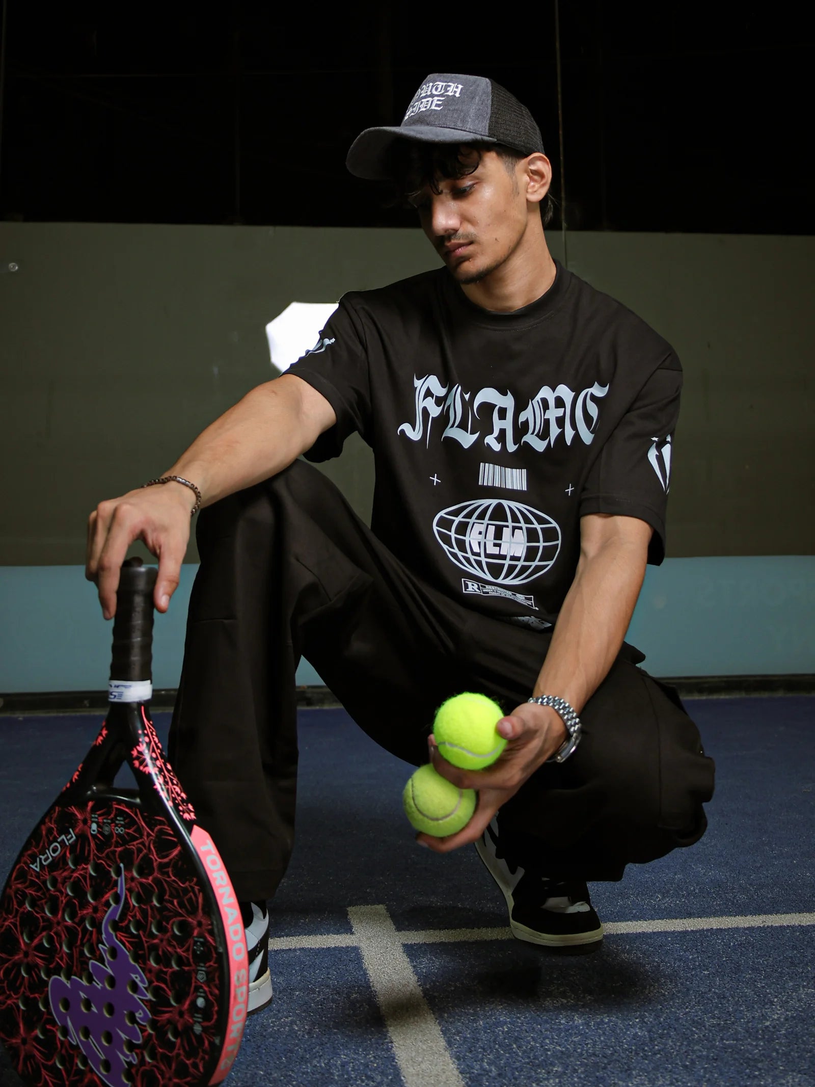 Person on a tennis court holding a racket and balls, wearing a black t-shirt with 'FLAME' branding.