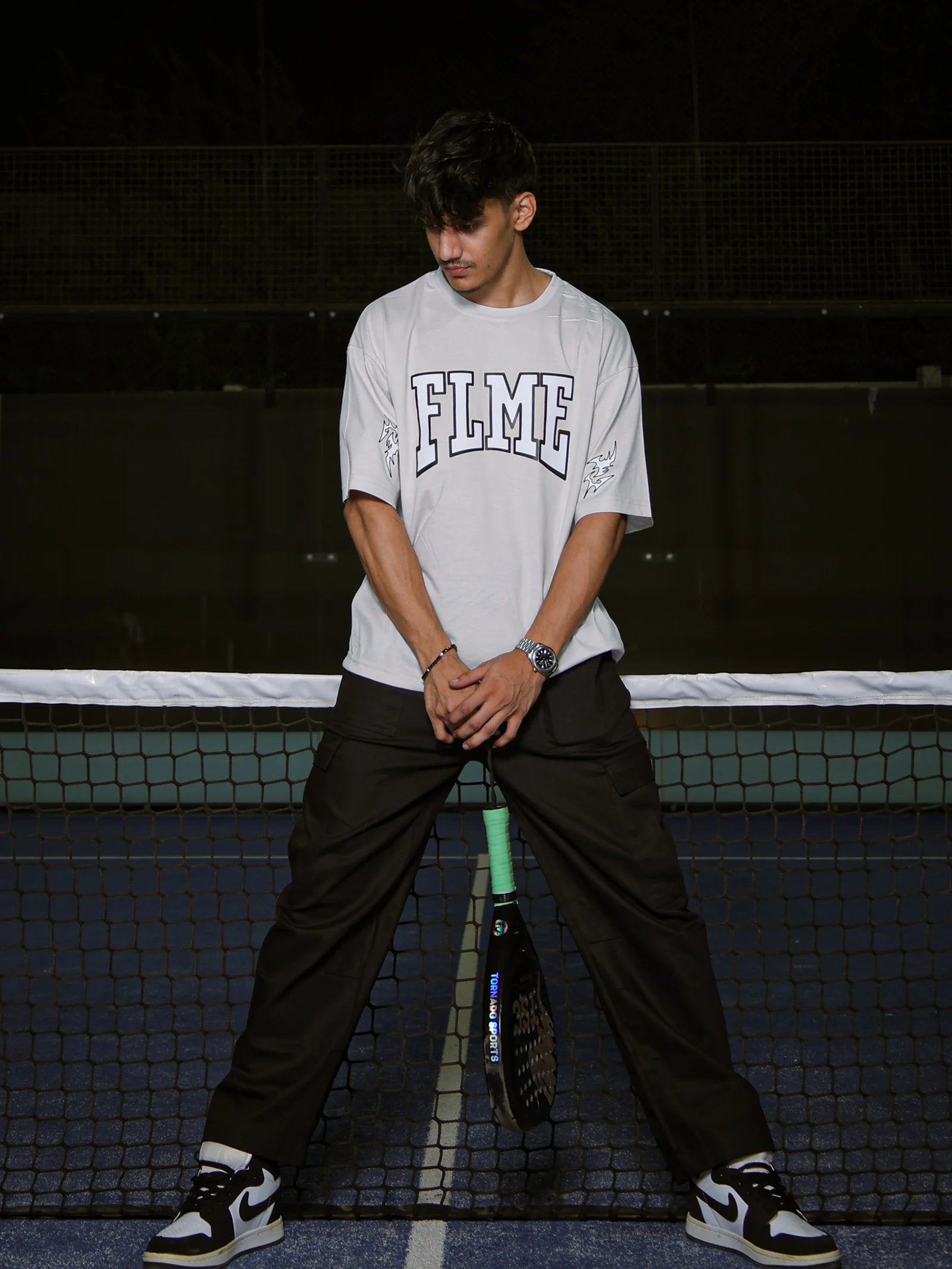 Person holding a tennis racket on a tennis court wearing a 'FLME' t-shirt.