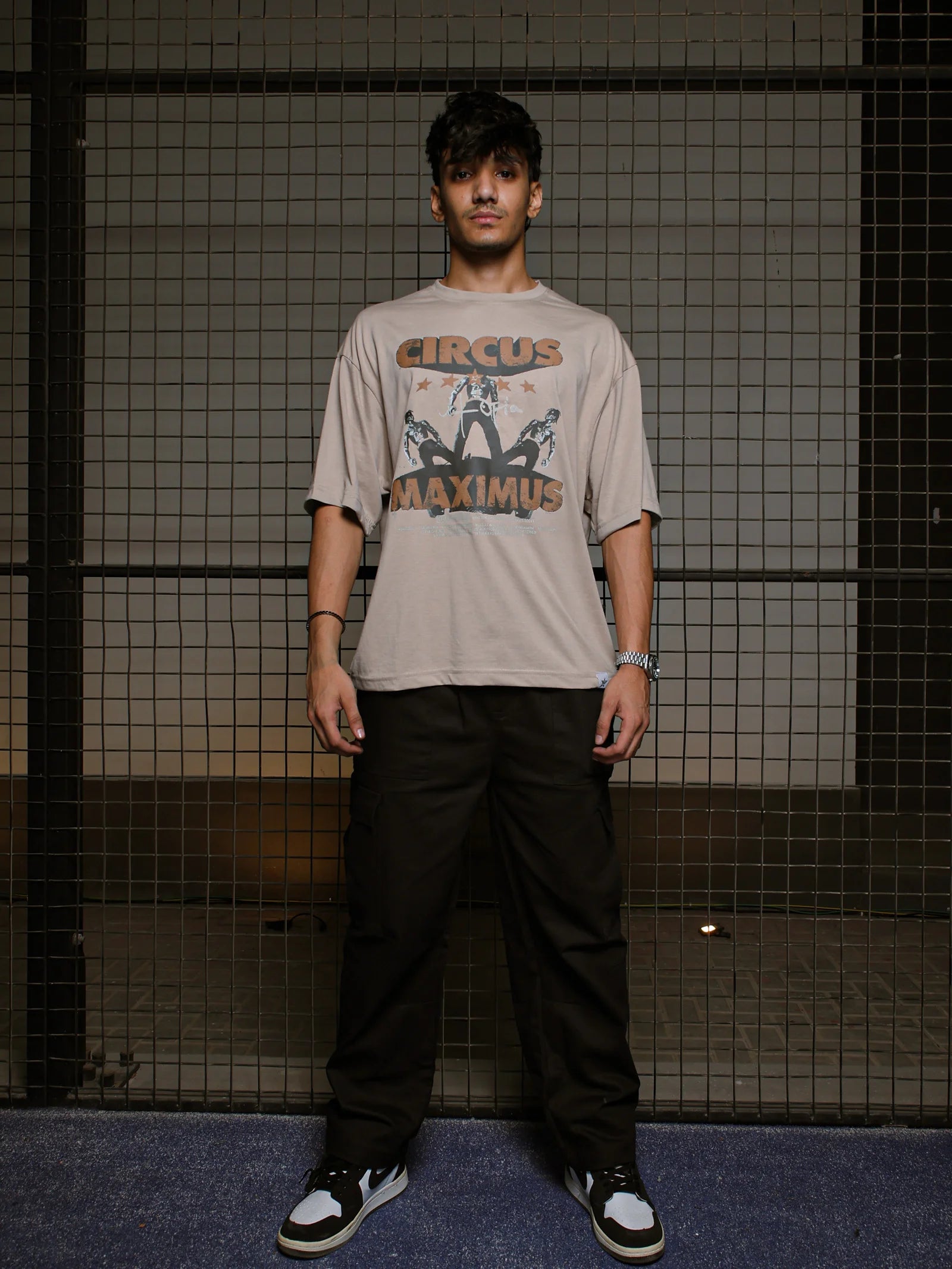 Person wearing a 'Circus Maximus' t-shirt standing against a grid-patterned wall.