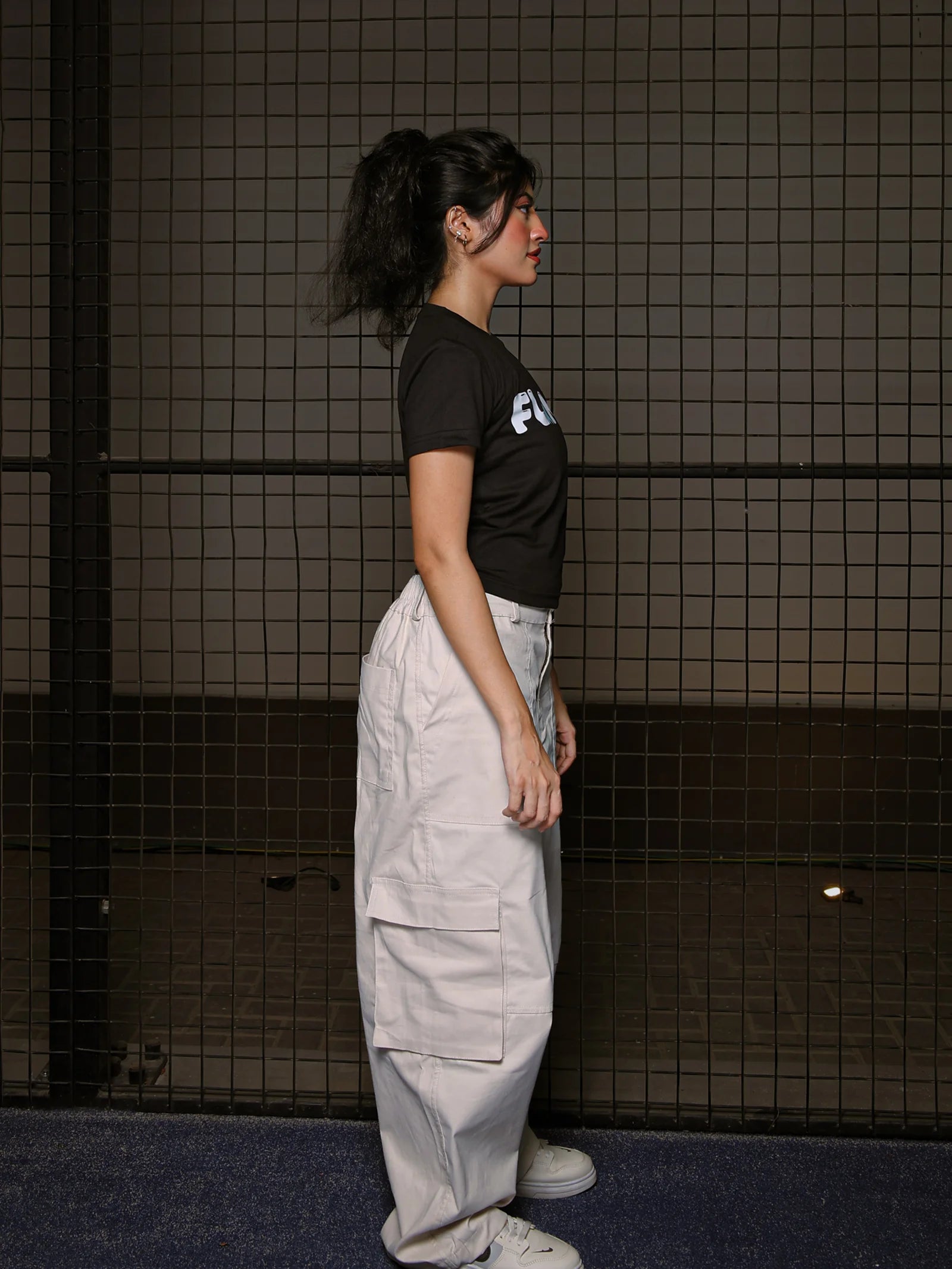 Person wearing a black t-shirt with a logo and light-colored pants standing against a tiled wall.