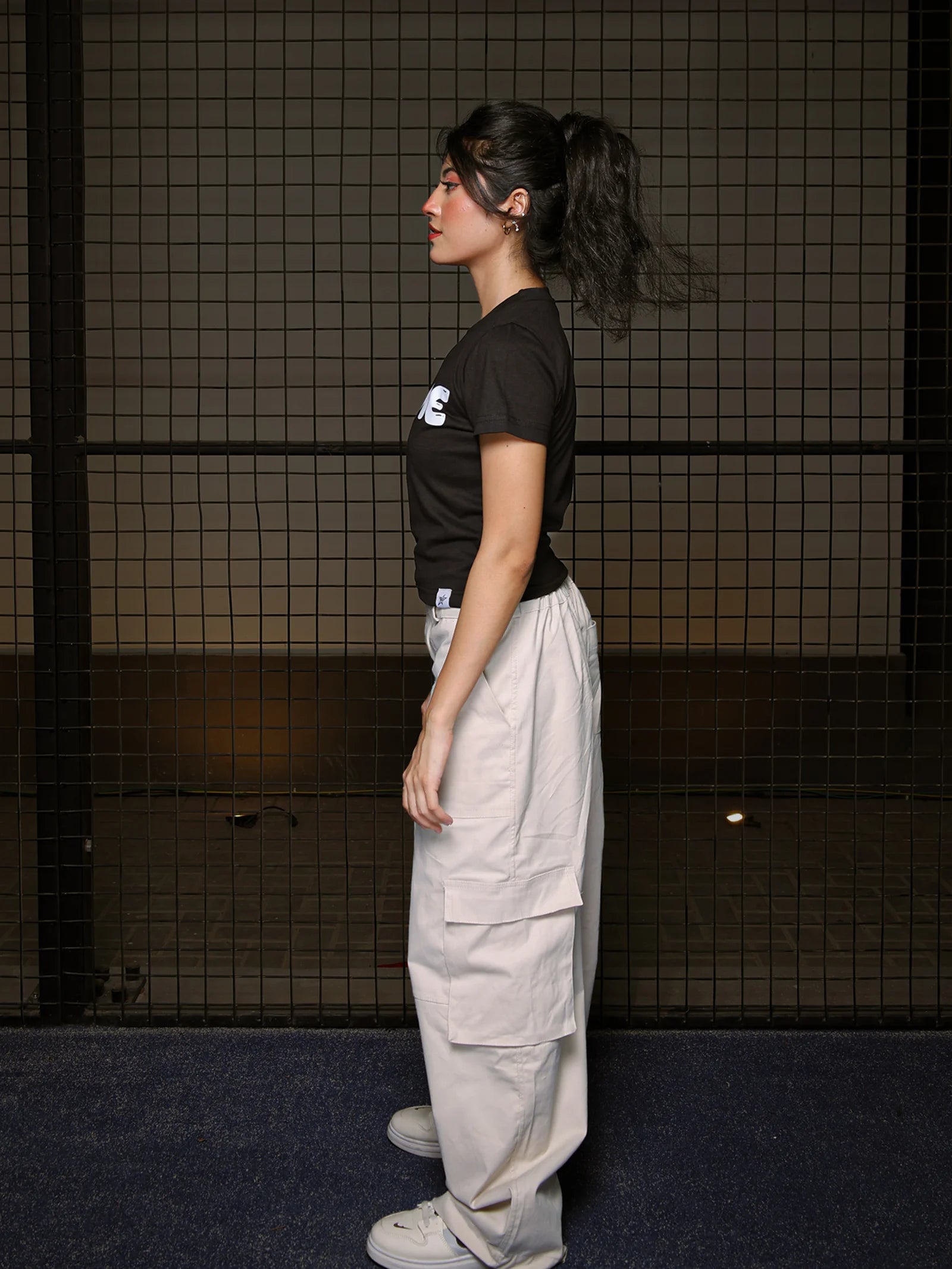 Person wearing a black t-shirt with a logo and light-colored cargo pants standing against a dark tiled wall.