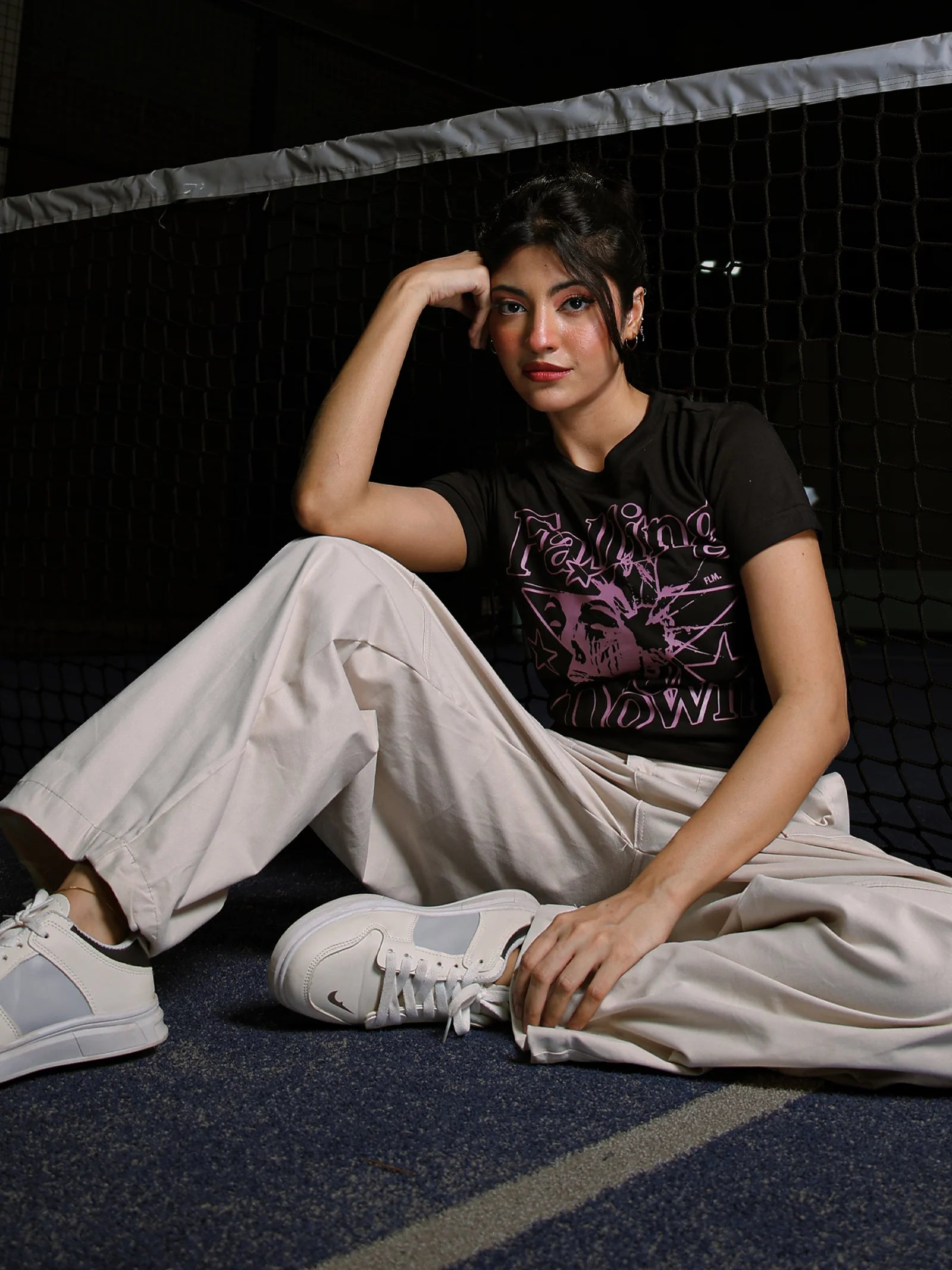 Person sitting on a dark floor wearing a black t-shirt with a graphic design and light-colored pants.