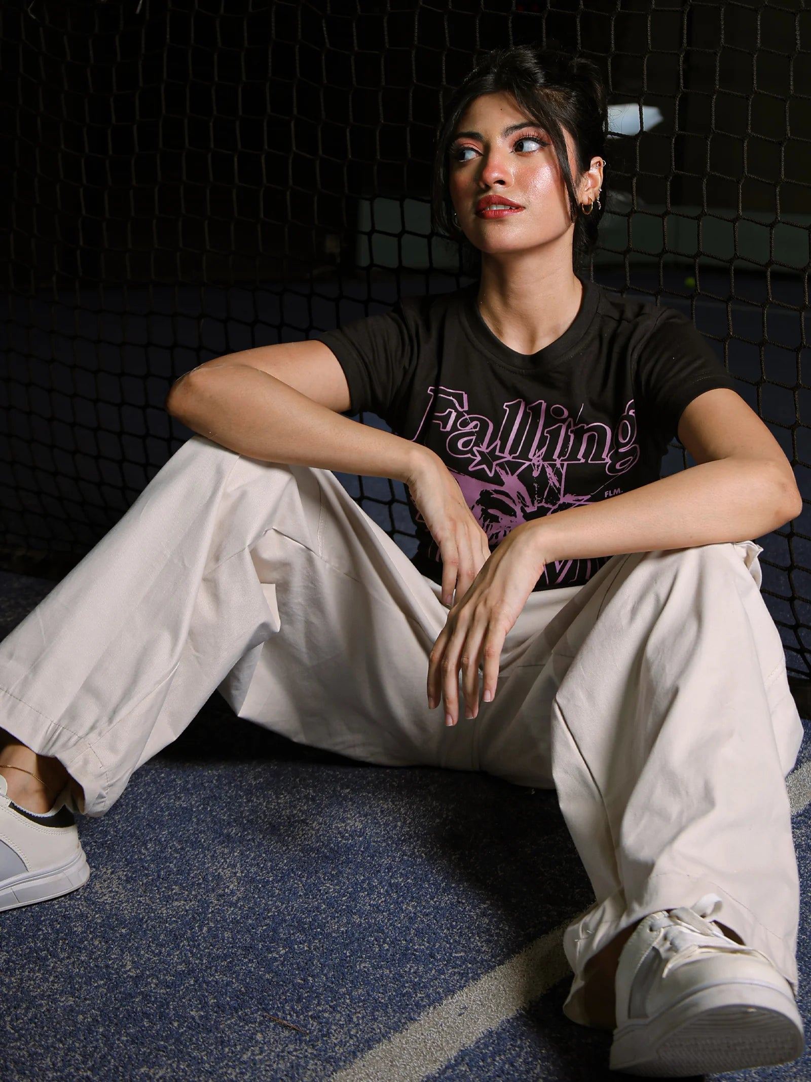 Person wearing a black t-shirt with text and light-colored pants sitting on a dark floor.