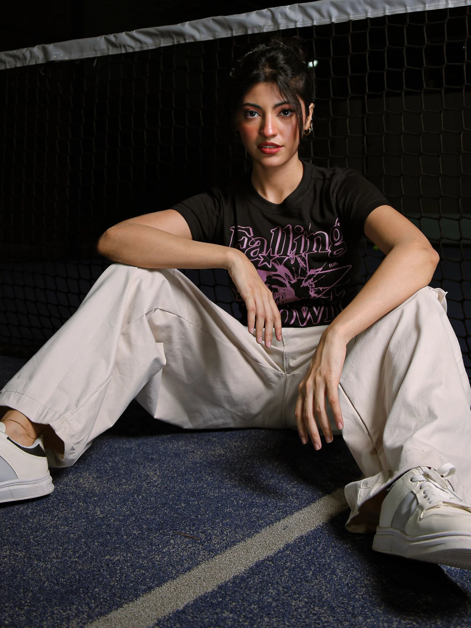 Person sitting on a dark floor wearing a black t-shirt with purple text and light-colored pants.