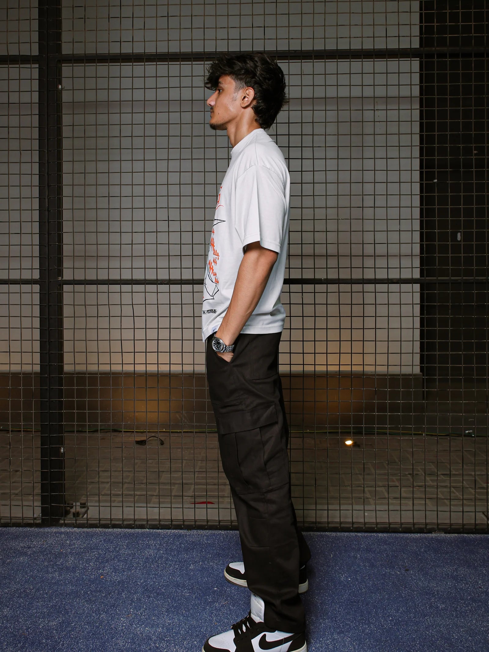 Person wearing a white t-shirt and black pants standing against a grid-patterned wall.