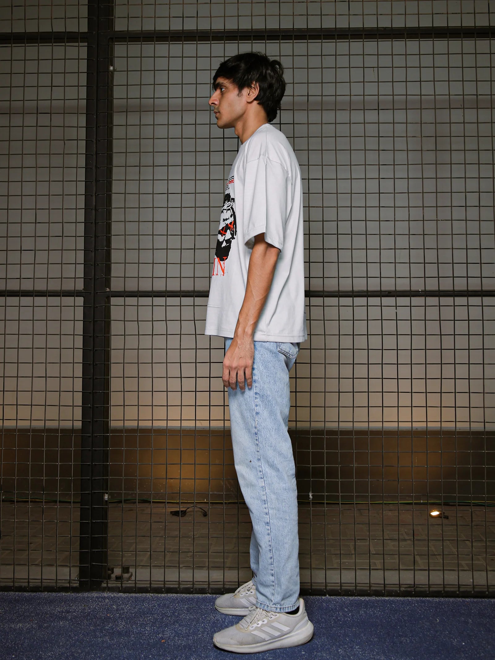 Person wearing a white t-shirt with a graphic design and light blue jeans standing against a tiled wall.