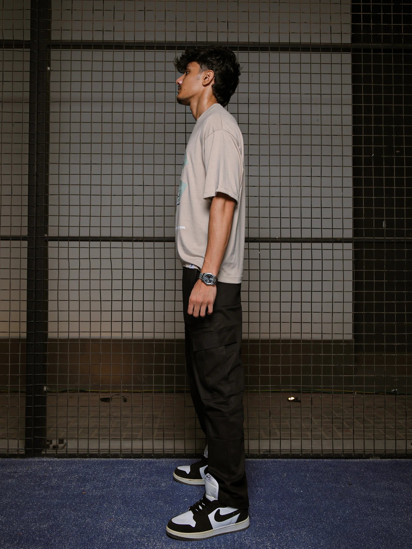 Man wearing a light gray t-shirt, black pants, and black and white sneakers against a tiled wall.