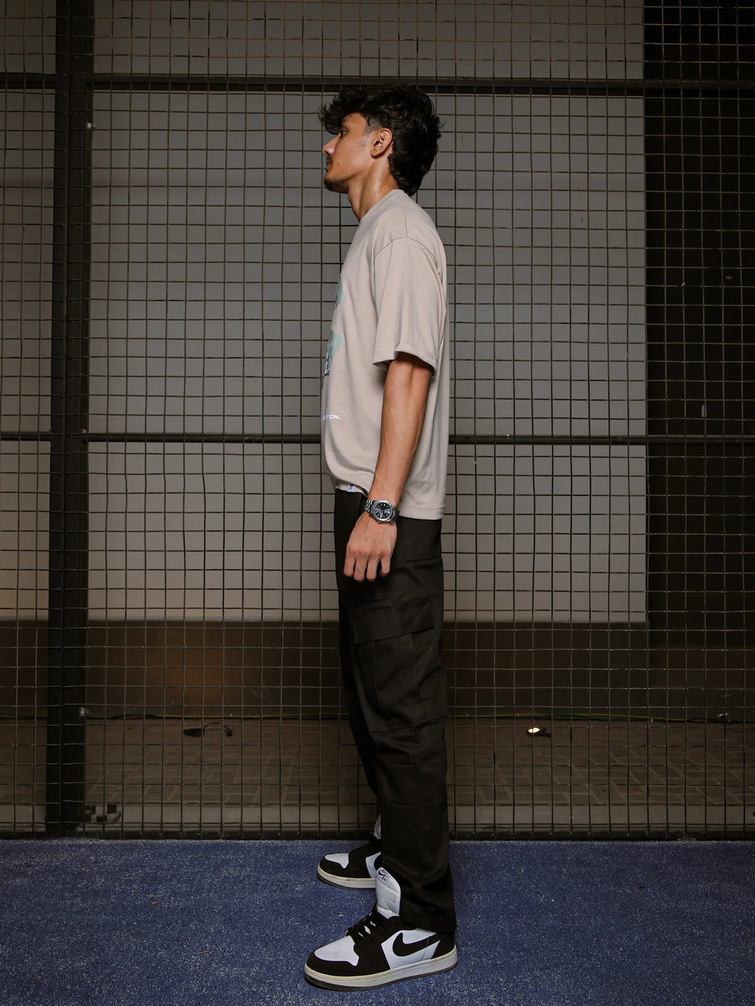 Man wearing a light gray t-shirt, black pants, and black and white sneakers against a tiled wall.