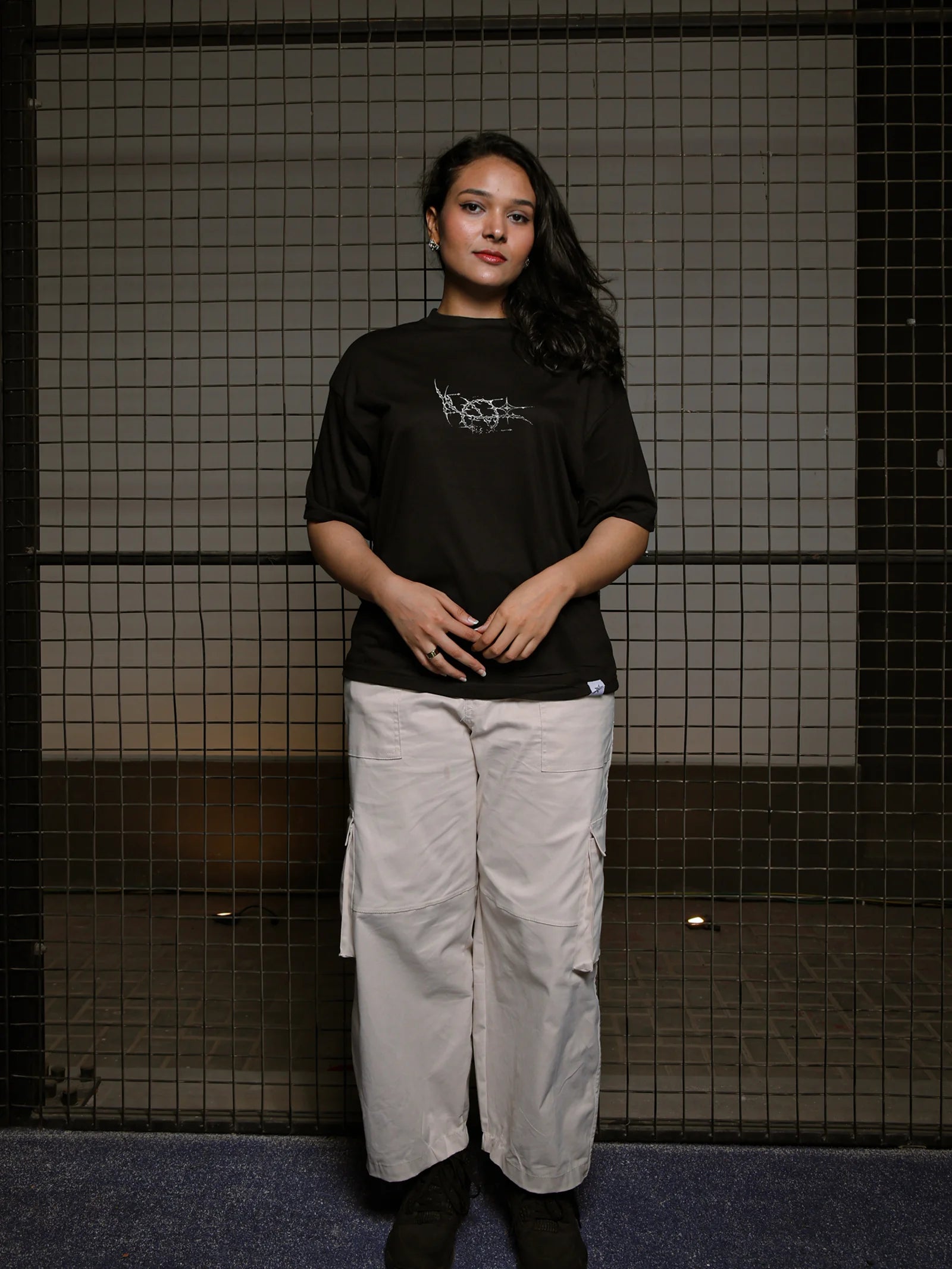 Person wearing a black t-shirt with a graphic design and light-colored pants standing against a tiled wall.