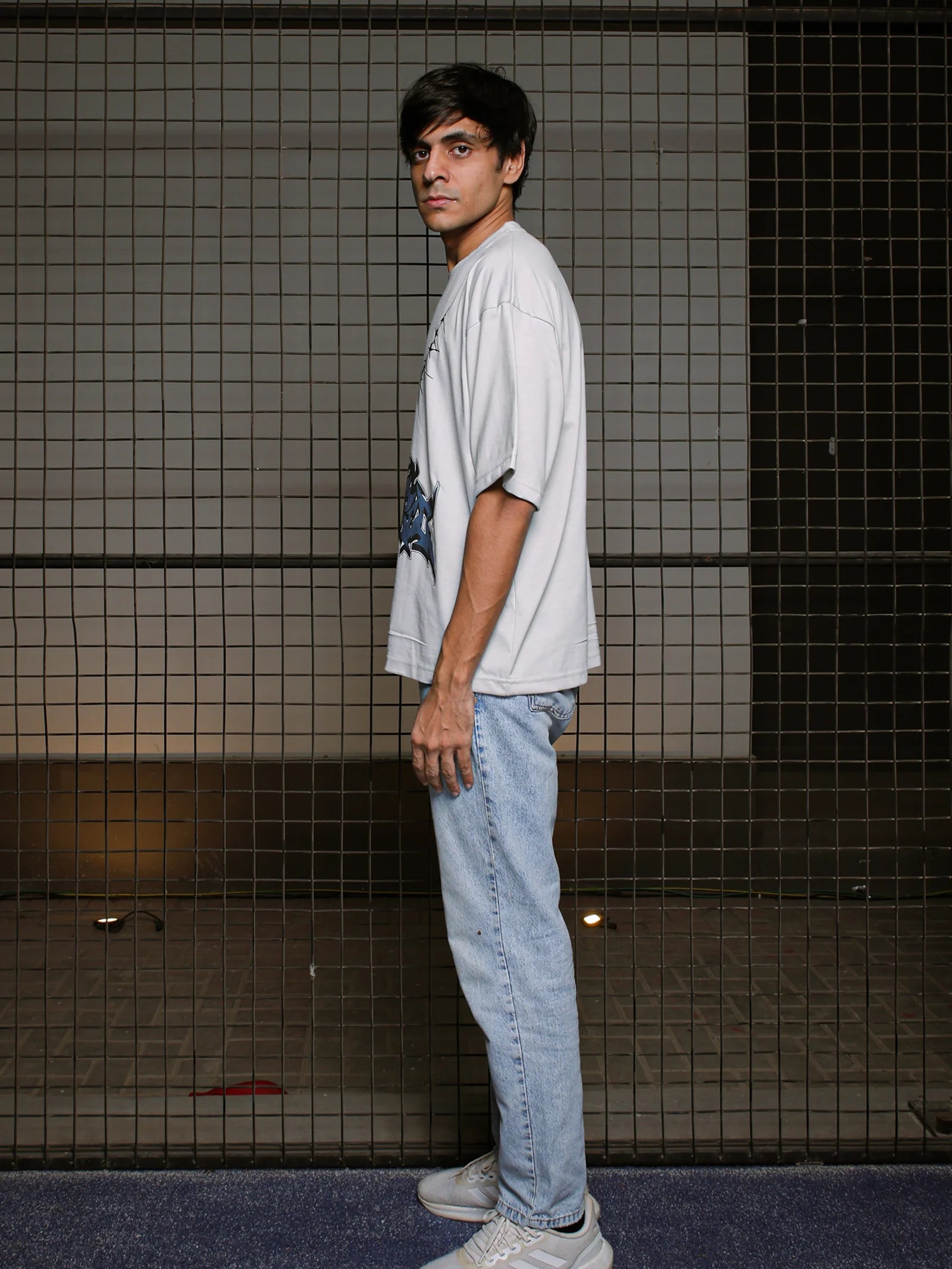Person wearing a white t-shirt and light blue jeans standing against a tiled wall.