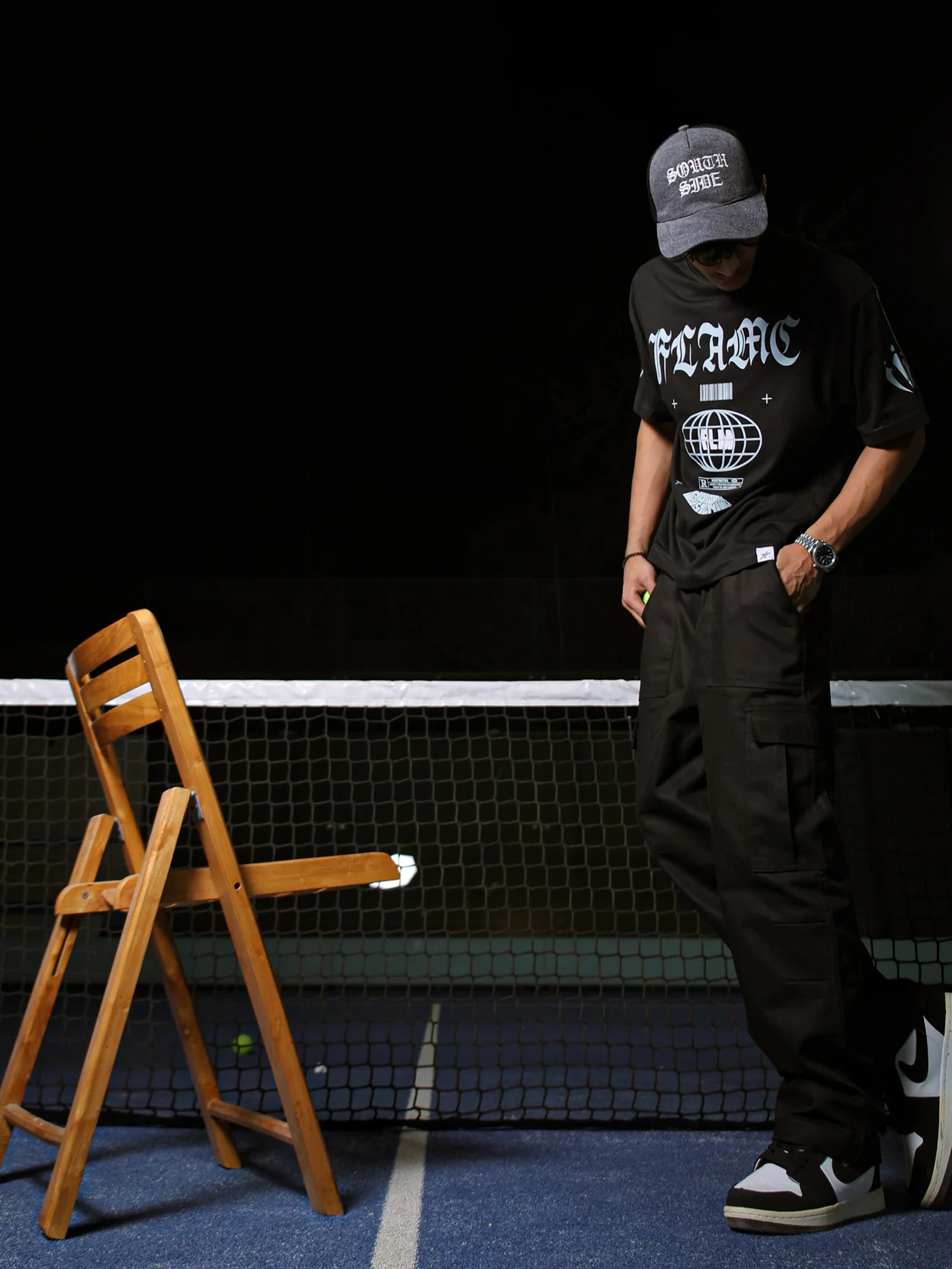 Person wearing a black t-shirt with white text and graphics, standing on a tennis court next to a wooden chair.