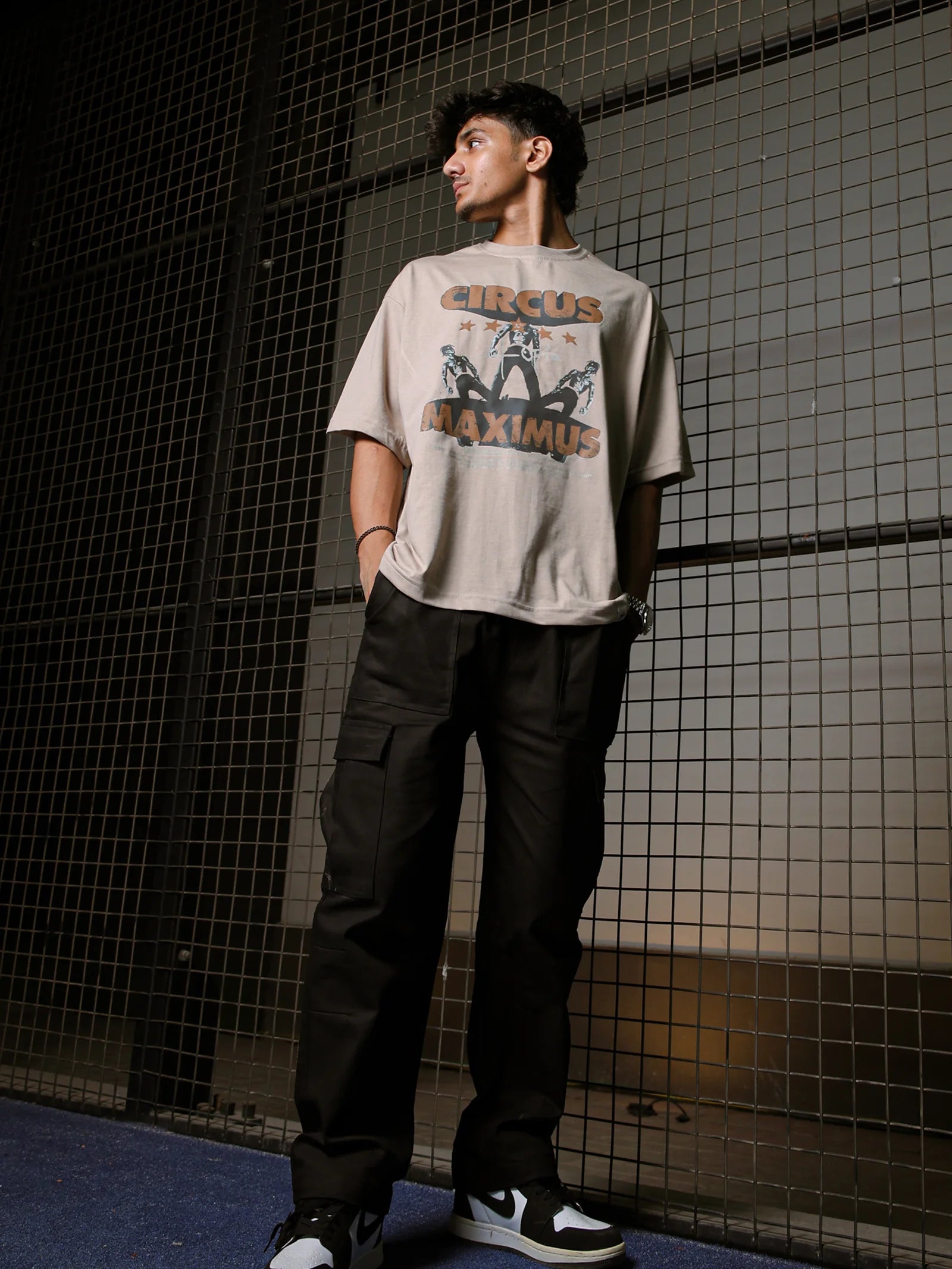 Person wearing a beige t-shirt with graphic design and text, standing against a grid-patterned wall.