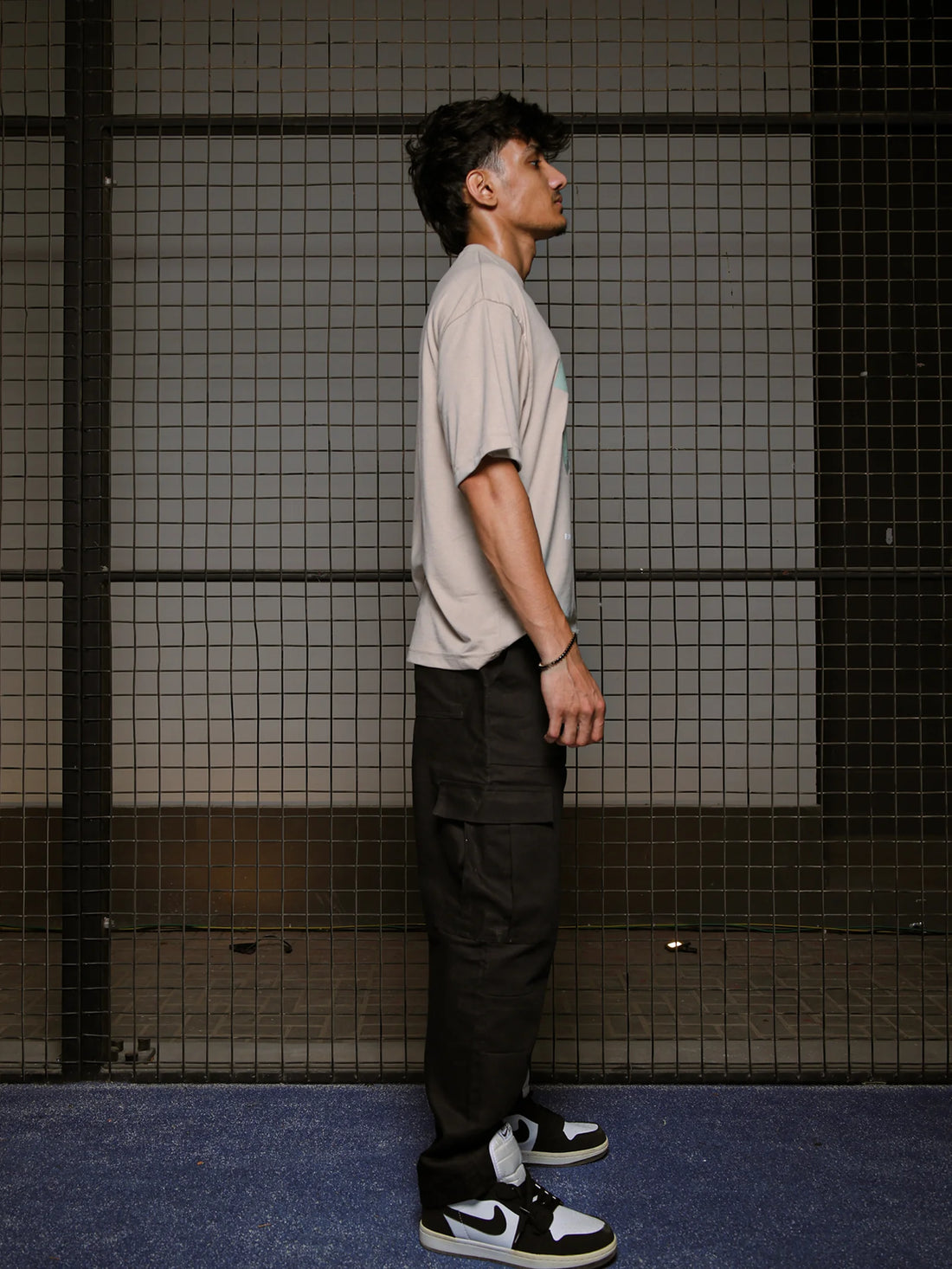 Person standing against a grid-patterned wall wearing a beige t-shirt and black pants.