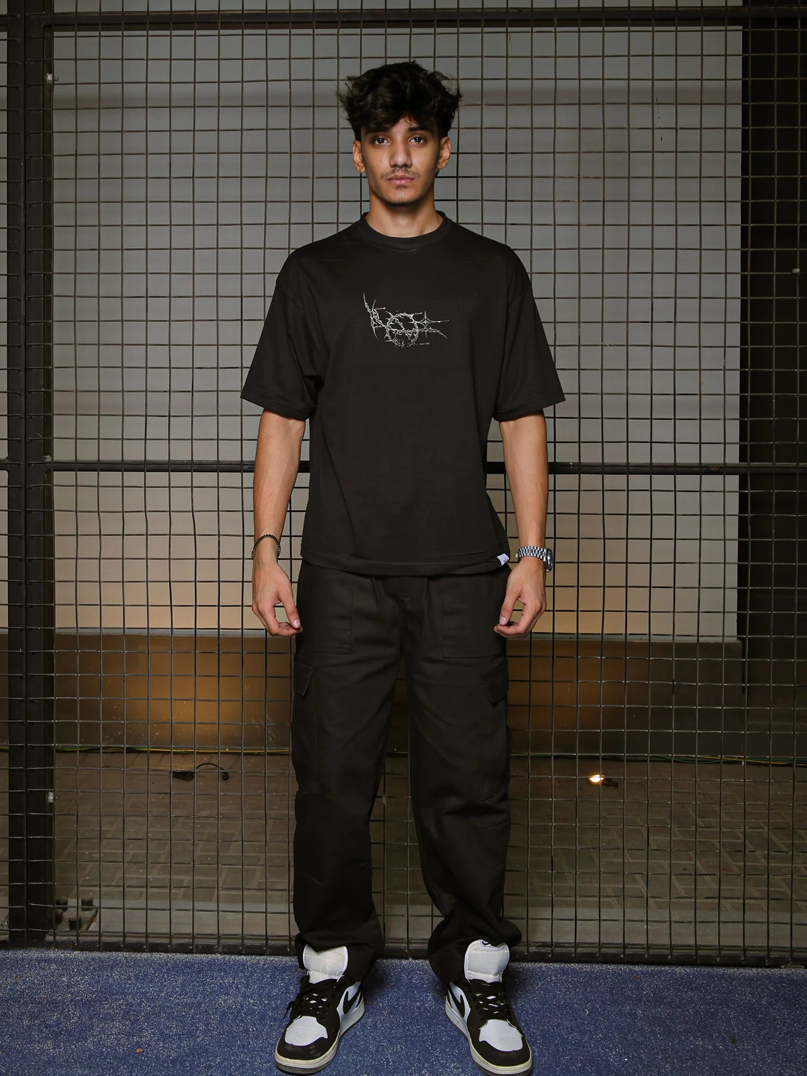 Person wearing a black t-shirt with a logo and black pants standing against a grid-patterned wall.