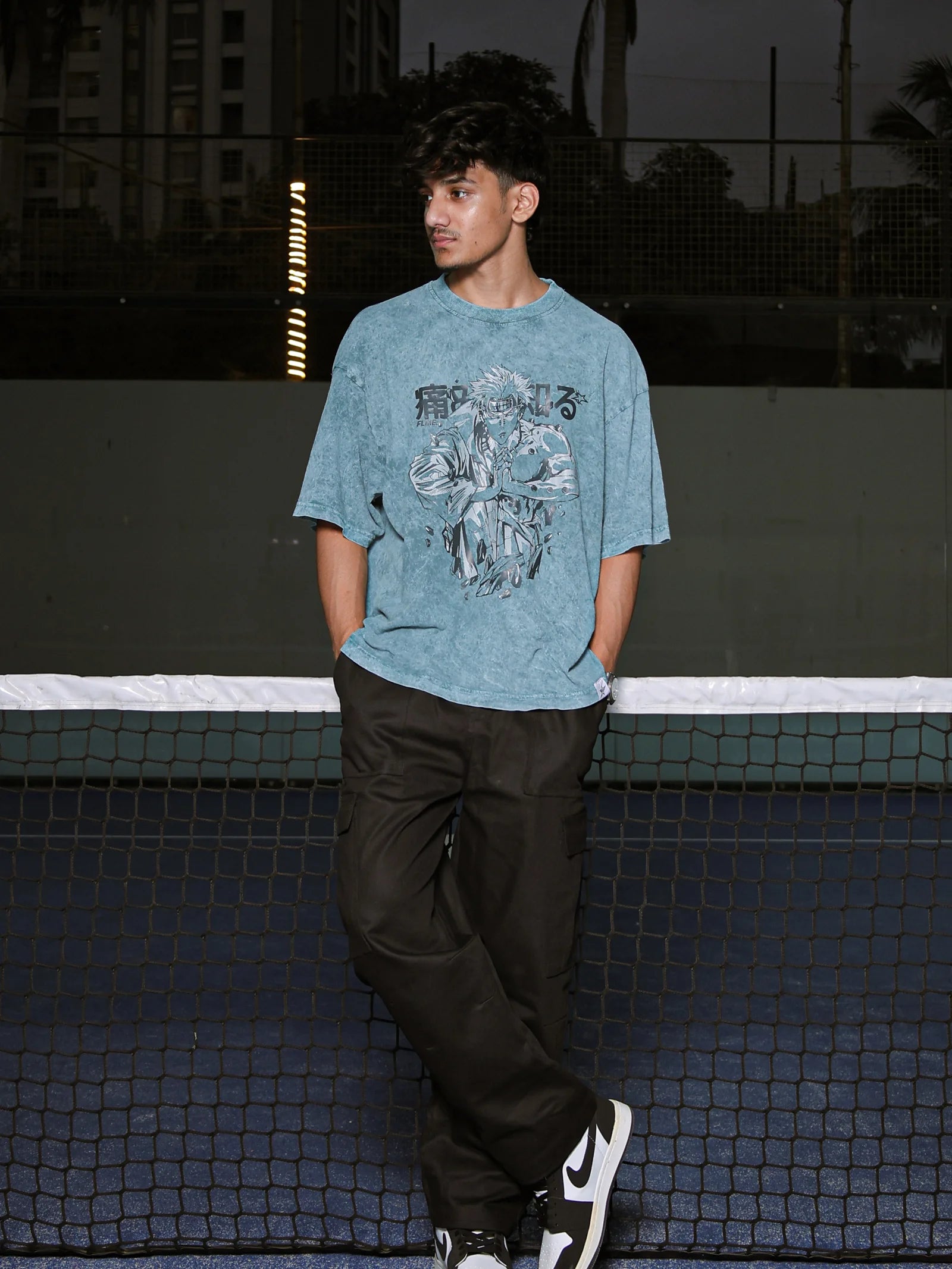 Person wearing a blue graphic t-shirt and black pants standing on a tennis court.