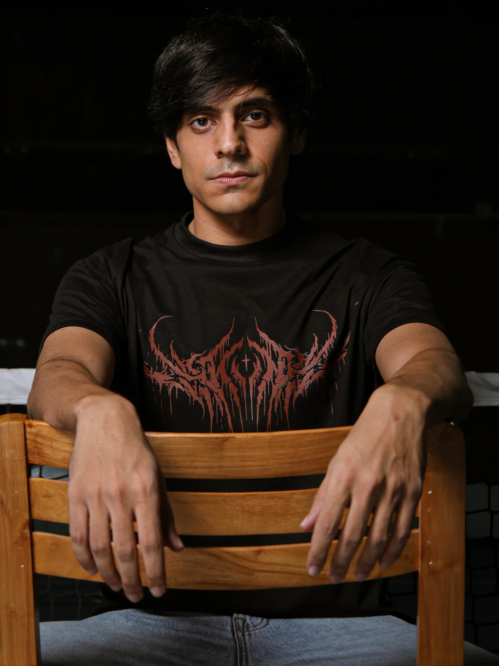 Man wearing a black t-shirt with a red graphic design, sitting on a wooden chair against a dark background.