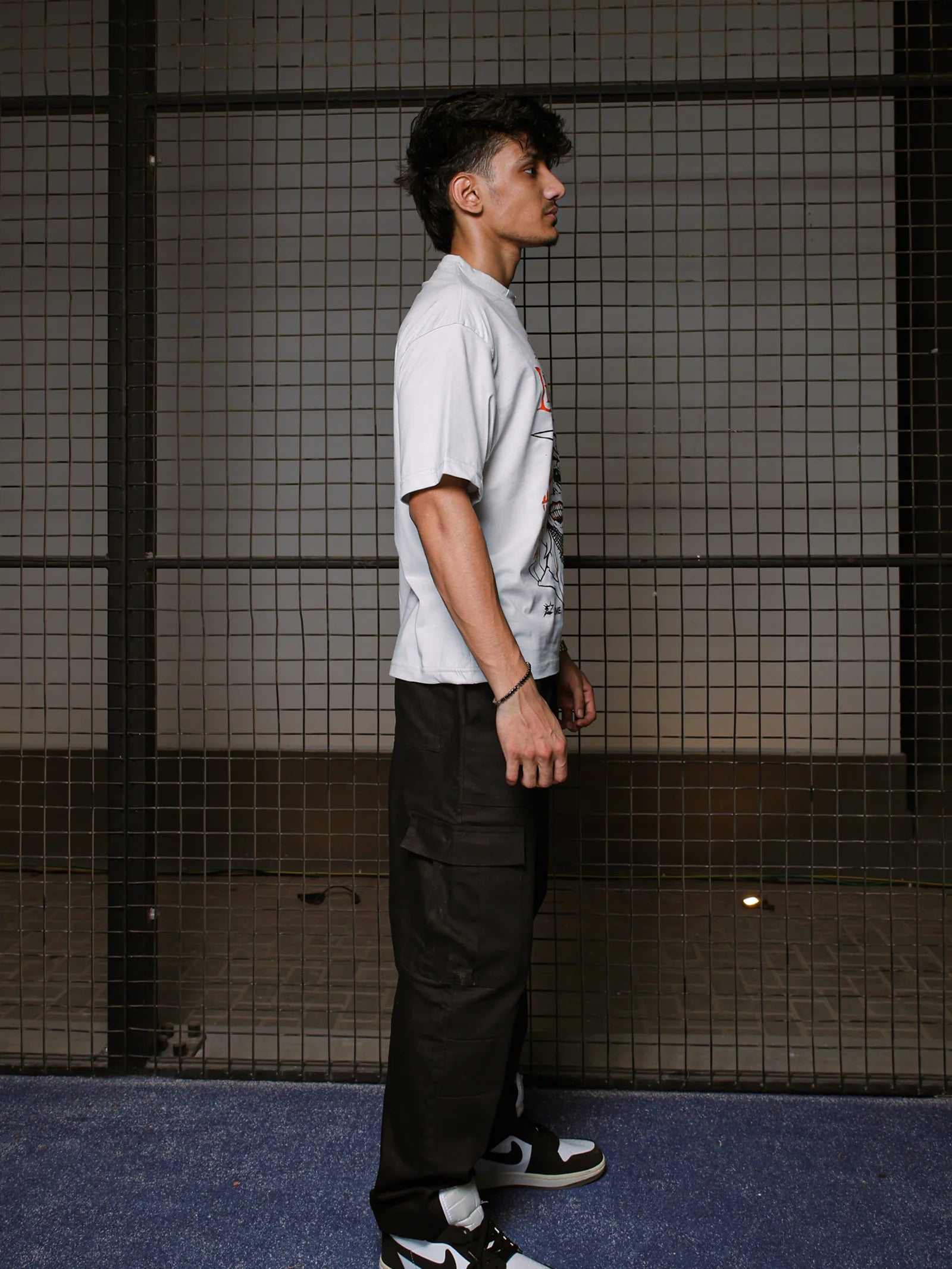 Person wearing a white t-shirt and black pants standing against a tiled wall.