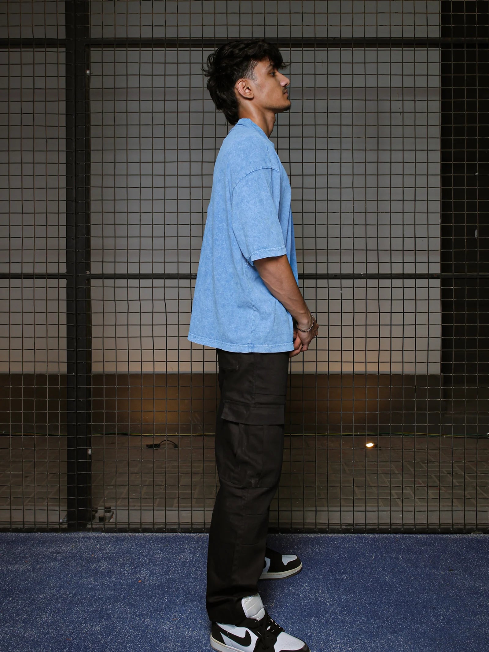 Person wearing a light blue shirt and black pants standing against a tiled wall.
