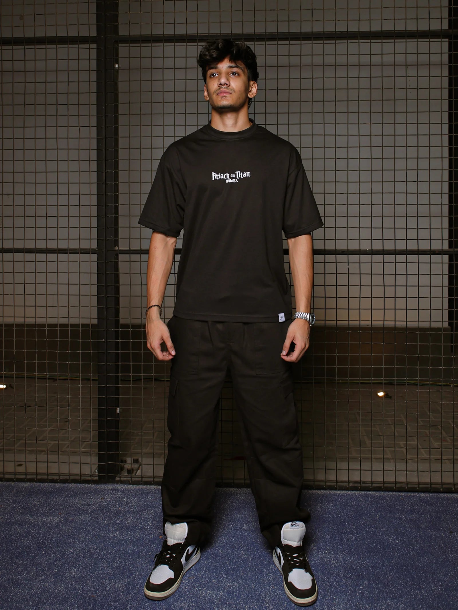Person wearing a black t-shirt with text and black pants standing against a grid-patterned wall.