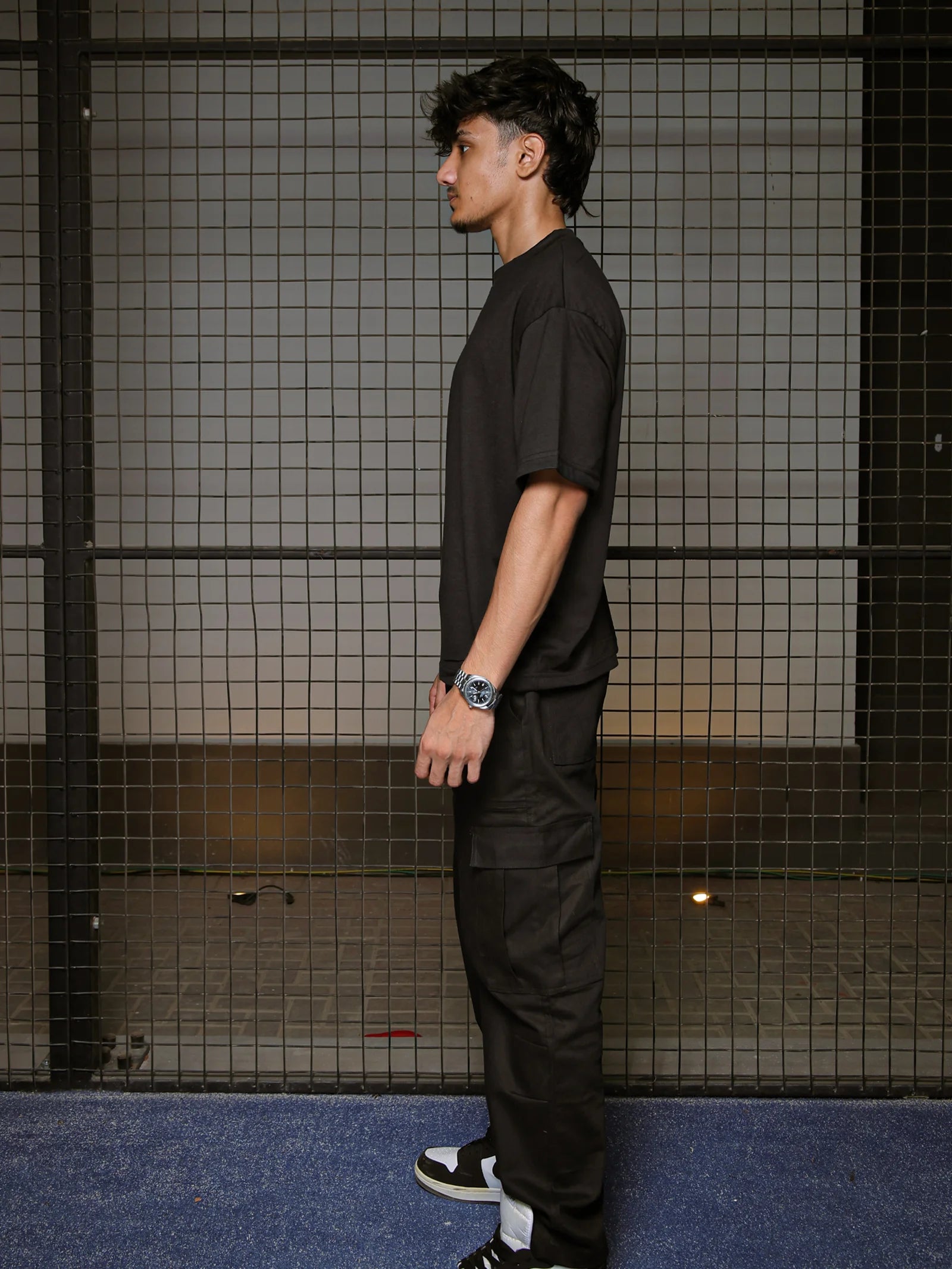 Man wearing a black outfit standing against a grid-patterned wall.