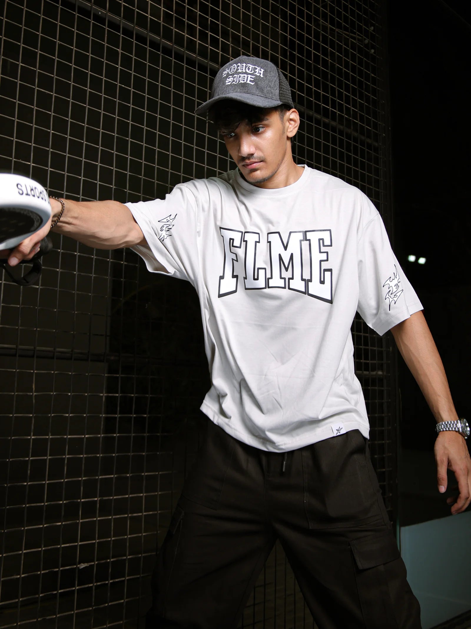 Person wearing a white t-shirt with 'ELME' branding and a cap, standing against a dark tiled wall.