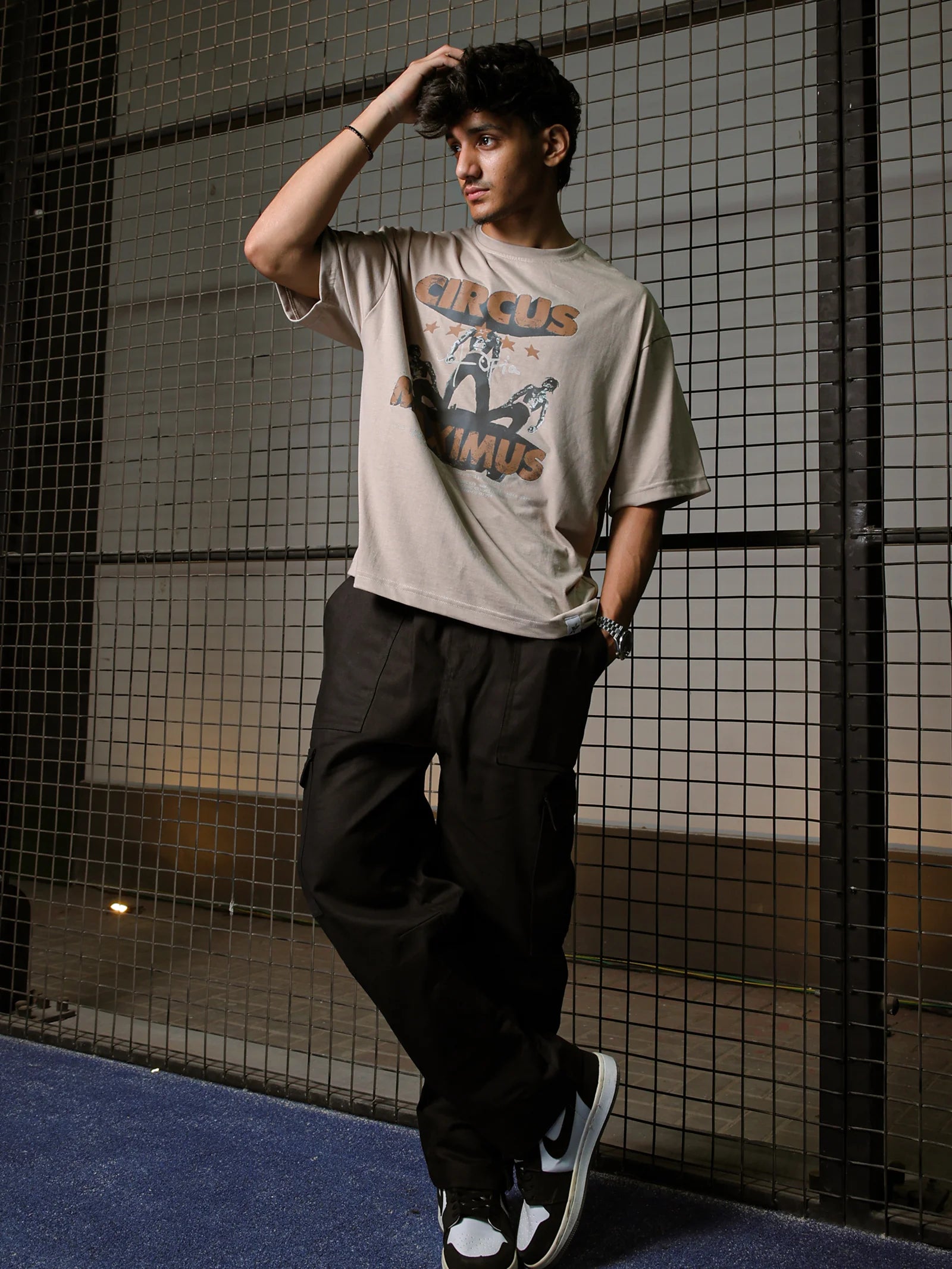 Person wearing a beige t-shirt with graphics and dark pants against a tiled wall.