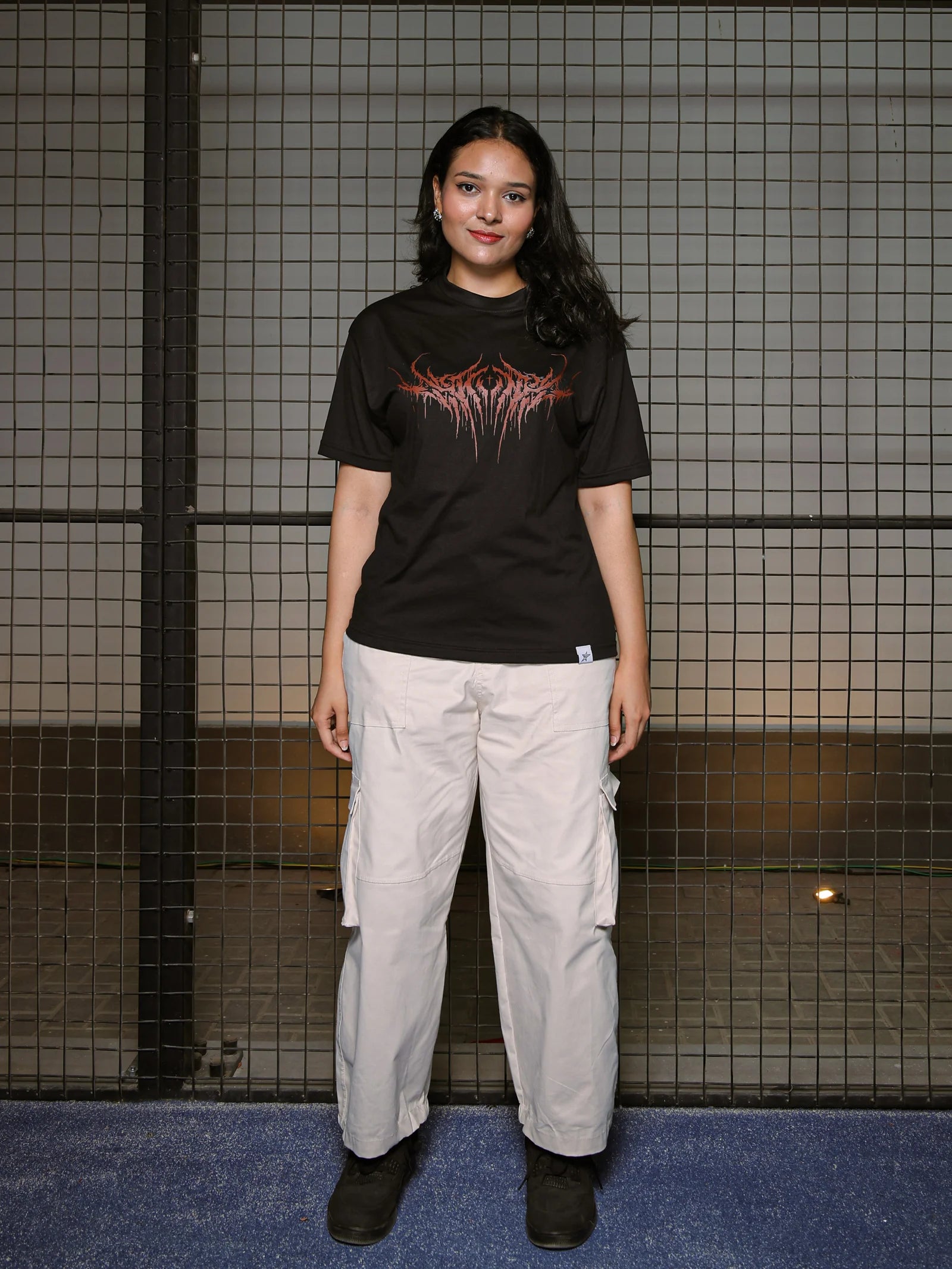 Person wearing a black t-shirt with red graphic design and light gray cargo pants standing against a tiled wall.