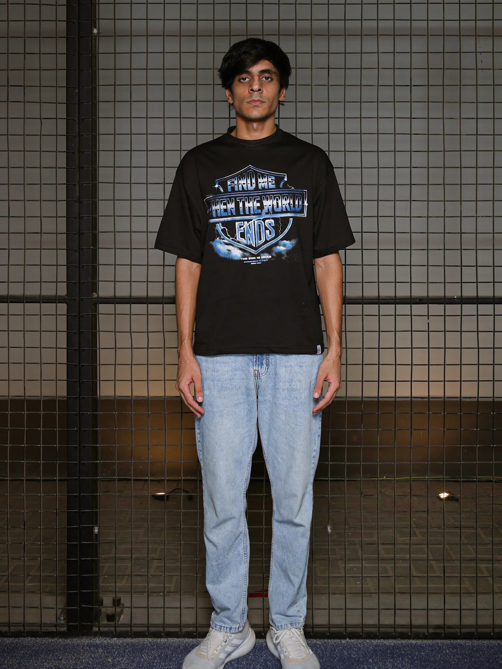 Person wearing a black t-shirt with a graphic design and light blue jeans standing against a tiled wall.