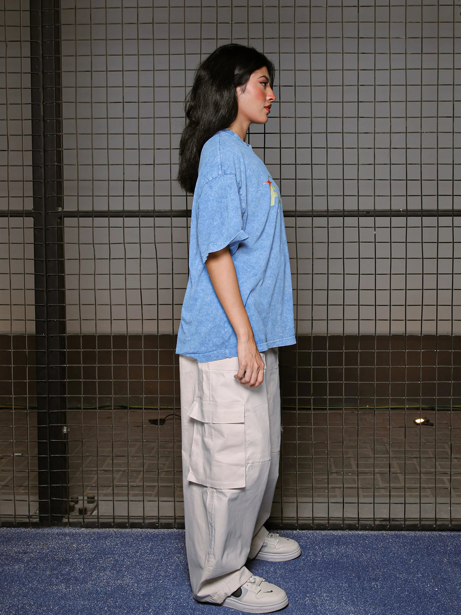 Person wearing a blue oversized shirt and beige cargo pants standing against a tiled wall.