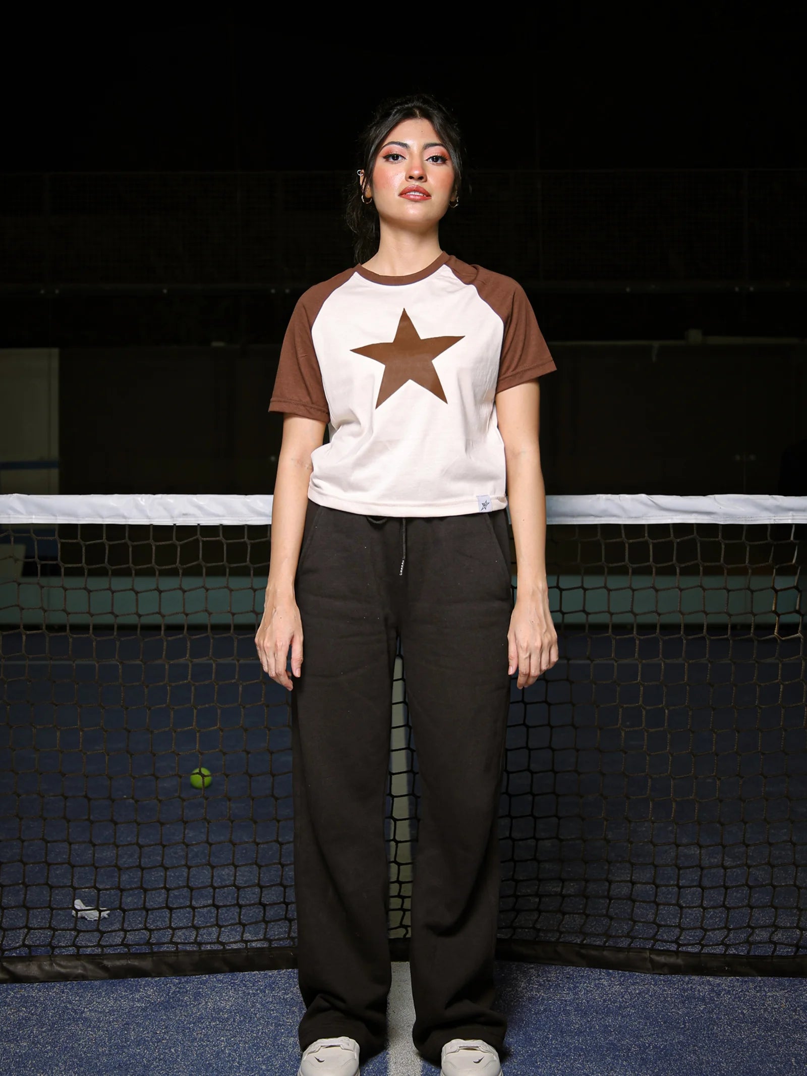 Person wearing a white and brown baseball shirt with a star design, standing in front of a tennis net.