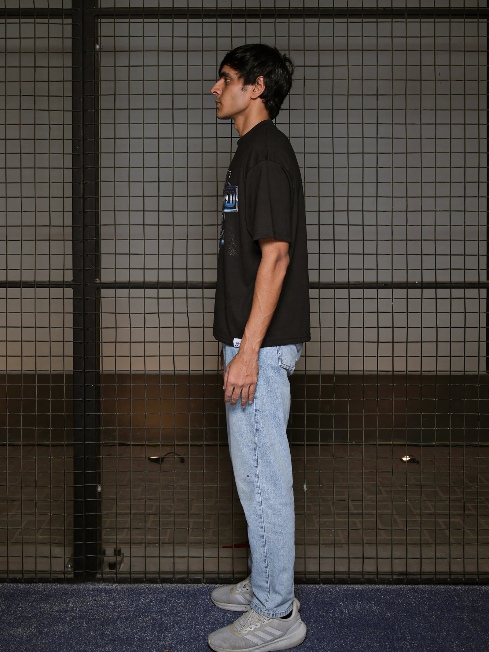 Person wearing a black shirt and light blue jeans standing in front of a tiled wall.