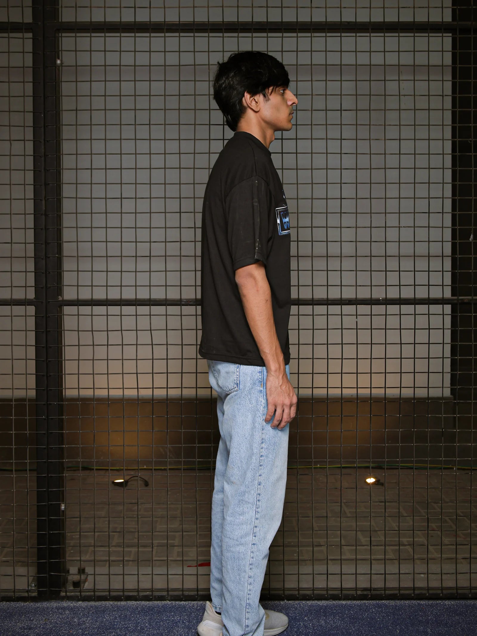 Person wearing a black t-shirt and light blue jeans standing against a tiled wall.